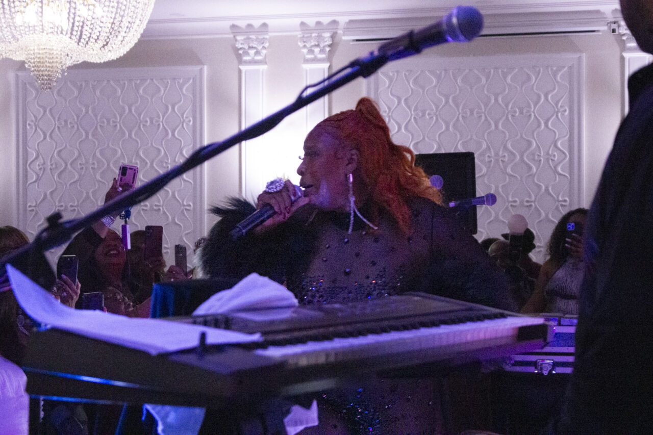 Performer Macka Diamond onstage, framed betwixt a microphone stand and a keyboard in the foreground